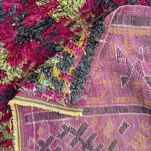 May include: A close-up of a handmade rug with a vibrant, textured design. The rug features a mix of colors, including deep pink, green, and gray, creating a visually rich pattern. The edges are finished with a fringe.