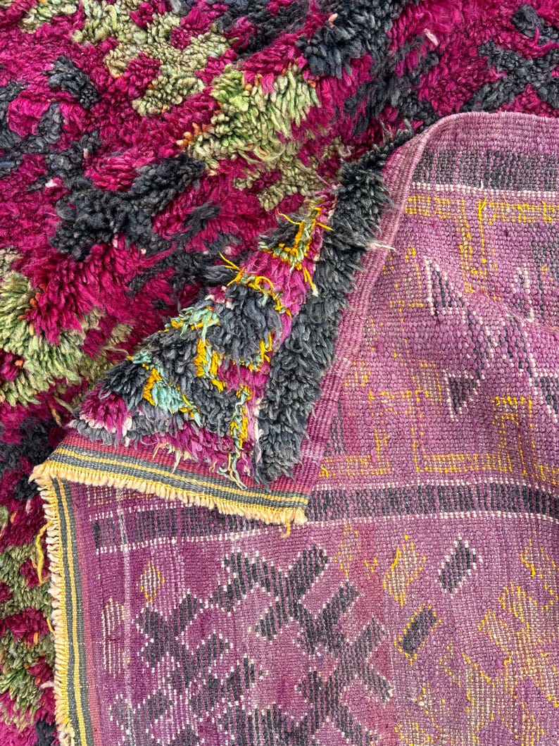 May include: A close-up of a handmade rug with a vibrant, textured design. The rug features a mix of colors, including deep pink, green, and gray, creating a visually rich pattern. The edges are finished with a fringe.