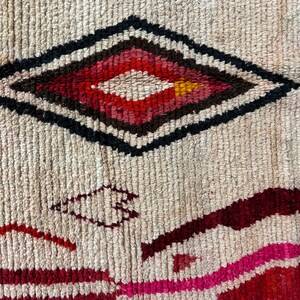 May include: A close-up of a woven rug with a diamond-shaped pattern in shades of red, pink, black, and white. The rug has a textured surface and a cream background.