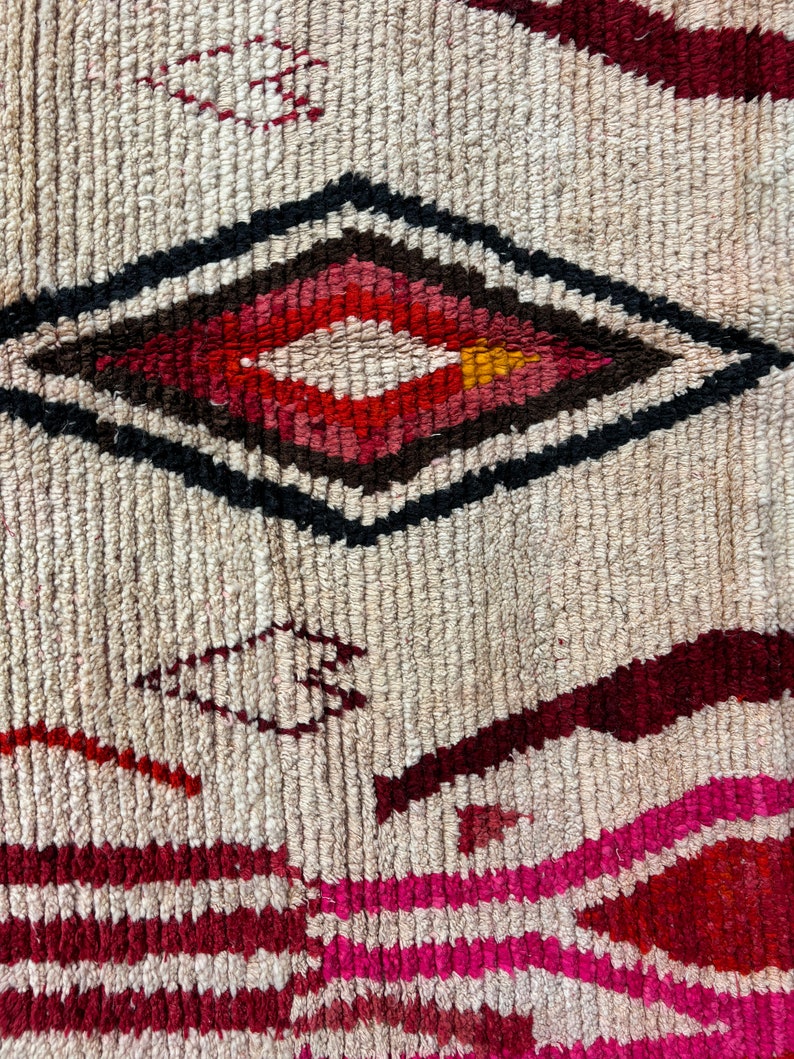 May include: A close-up of a woven rug with a diamond-shaped pattern in shades of red, pink, black, and white. The rug has a textured surface and a cream background.