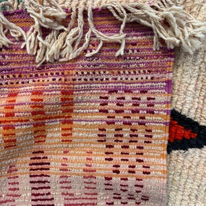 May include: A close-up of a woven rug with a colourful geometric pattern. The rug has a fringe border and is made of natural fibres.