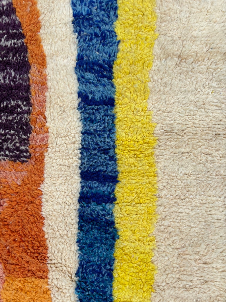 May include: Close-up of a colorful striped rug with a white, blue, yellow, and orange pattern. The rug is made of a soft, fluffy material.