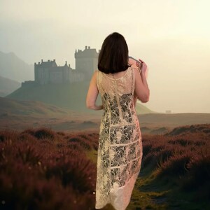 May include: A woman wearing a white lace dress stands in a field, with a castle in the distance. The field is covered in purple heather and the sky is a soft pink.