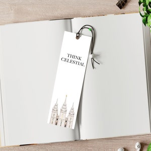 May include: A white bookmark with the text "THINK CELESTIAL" and an illustration of a building. The bookmark is placed in an open book on a wooden surface, with a coffee cup, earbuds, and greenery nearby.