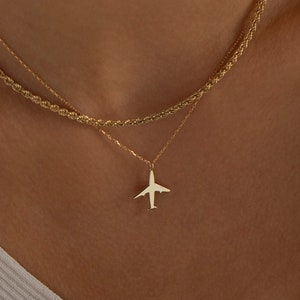May include: A gold chain necklace with a small airplane pendant. The necklace is layered on top of a thicker gold chain.