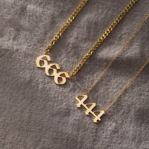 May include: Two gold necklaces with pendant charms. The top necklace has a pendant charm with the number 666 in a gothic font. The bottom necklace has a pendant charm with the number 444 in a gothic font.
