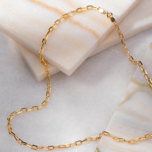 May include: A gold chain necklace with a delicate, square-shaped link design. The chain is laying on a white marble surface.