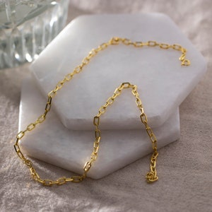 May include: A gold chain necklace with a delicate, square-shaped link design. The chain is lying on a white marble surface.