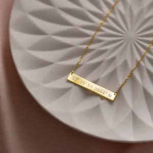 May include: A gold necklace with a bar pendant engraved with the coordinates "37&deg; 35' 51.2844" N. The necklace is on a white plate with a geometric pattern.