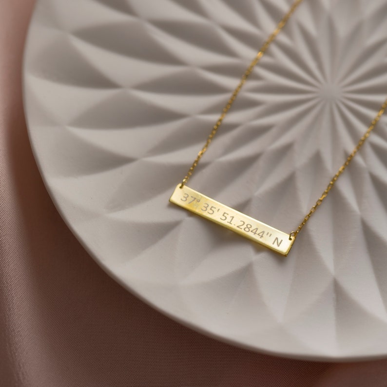 May include: A gold necklace with a bar pendant engraved with the coordinates "37&deg; 35' 51.2844" N. The necklace is on a white plate with a geometric pattern.