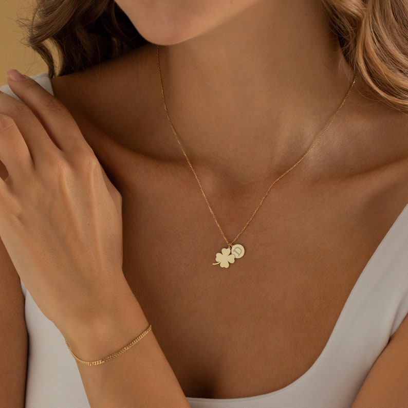 May include: A gold necklace with a four-leaf clover charm and a small round charm with the letter D engraved on it.