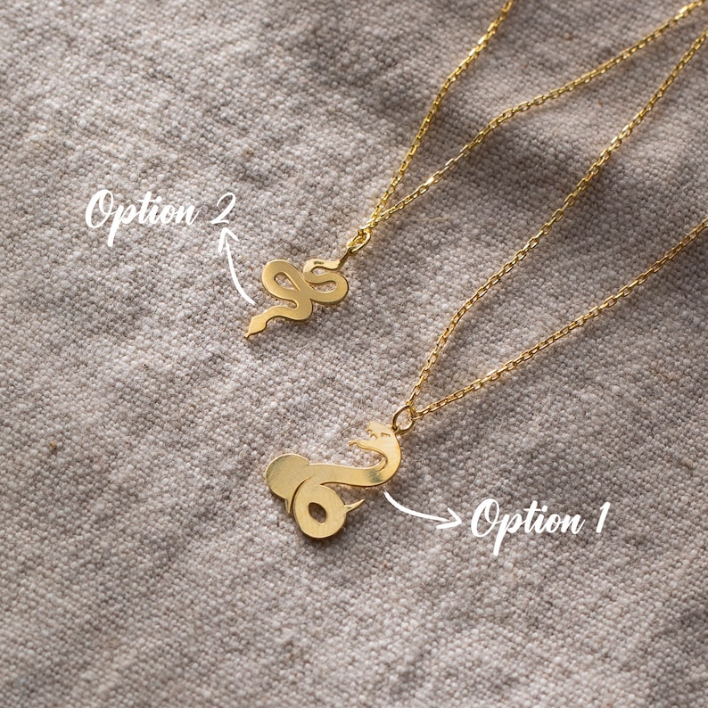 May include: Two gold-colored snake pendant necklaces. One necklace features a coiled snake design, while the other has a more stylized snake shape. The necklaces are displayed on a textured, neutral-toned surface.