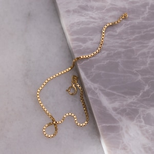 May include: A gold chain necklace with a box chain style. The chain is lying on a marble surface.