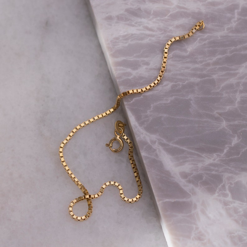 May include: A gold chain necklace with a box chain style. The chain is lying on a marble surface.