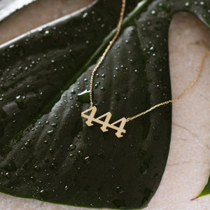 May include: Gold necklace with a pendant featuring the number 444. The necklace is laying on a green leaf with water droplets.