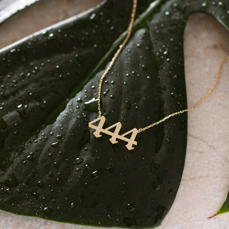 May include: Gold necklace with a pendant featuring the number 444. The necklace is laying on a green leaf with water droplets.