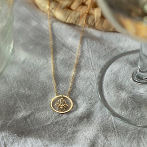 Puede incluir: Un collar de oro con un colgante de mano de Hamsa. El colgante es un c&iacute;rculo con un recorte de mano de Hamsa y un ojo en el centro.