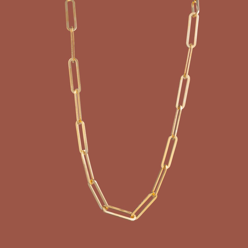 May include: A gold-colored chain necklace with elongated rectangular links. The necklace hangs against a solid brown background, showcasing its simple yet elegant design. The chain is made of a shiny, metallic material.