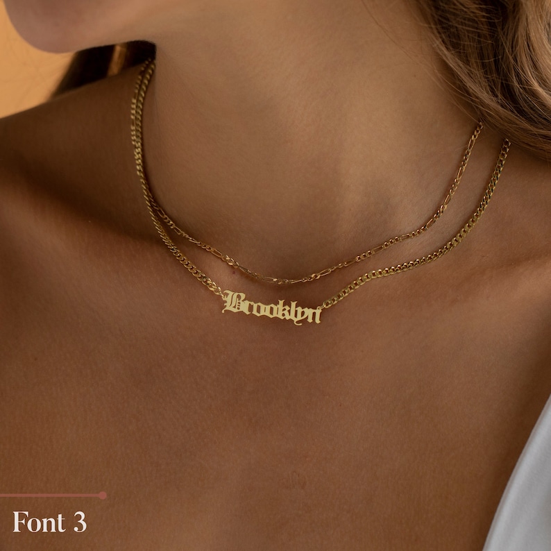 May include: A gold chain necklace with a gold nameplate pendant that says "Brooklyn" in a gothic font.