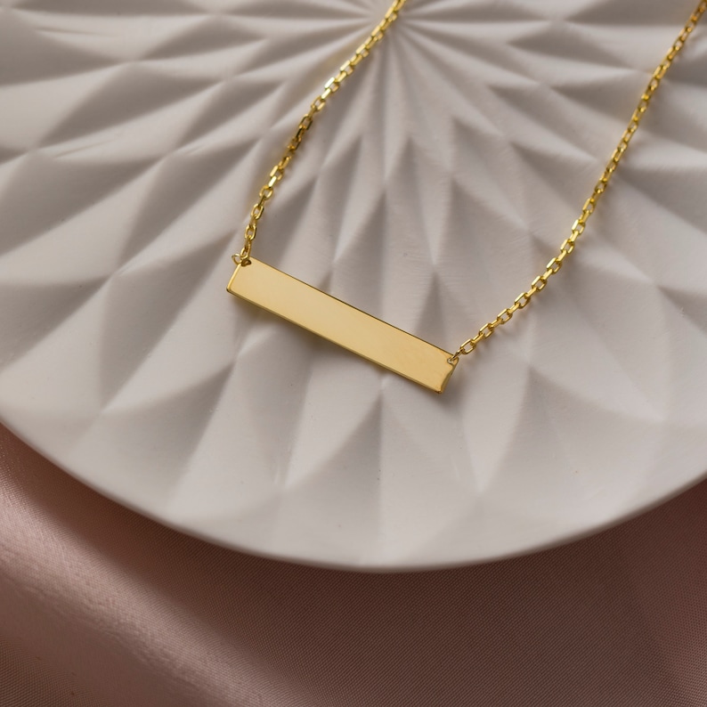 May include: A gold bar necklace with a delicate chain. The bar pendant is smooth and has a polished finish.