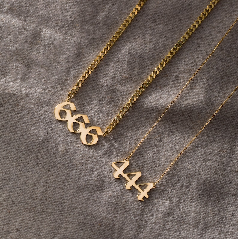 May include: Two gold necklaces with pendant charms. The top necklace has a pendant charm with the number 666 in a gothic font. The bottom necklace has a pendant charm with the number 444 in a gothic font.