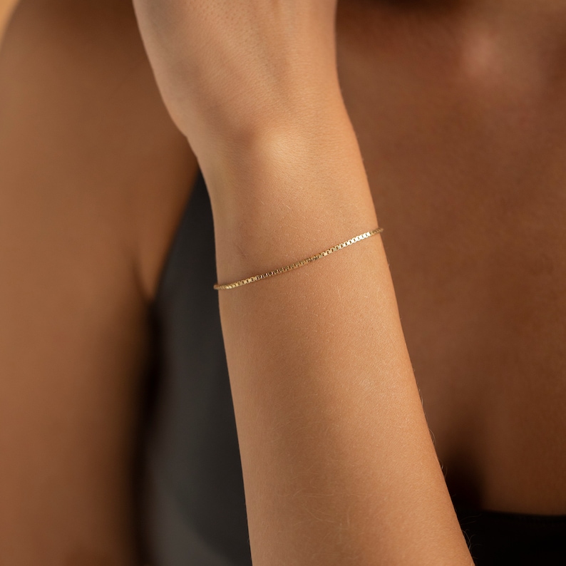 May include: A delicate gold chain bracelet with a box chain design. The bracelet is worn on a person's wrist.