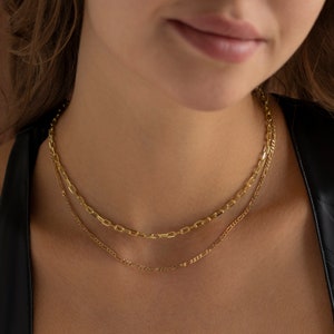 May include: Two gold chain necklaces layered on a person's neck. The top chain is a thicker, box chain and the bottom chain is a thinner, figaro chain.