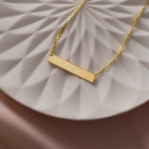 May include: A gold bar necklace with a delicate chain. The bar pendant is smooth and has a polished finish.