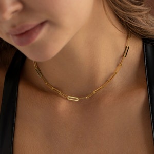 May include: A gold-colored paperclip chain necklace. The necklace is made of elongated, rectangular links. The chain is worn around the neck. The background is a blurred view of a person's skin and clothing.
