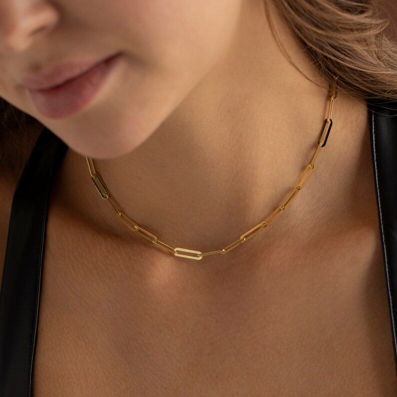 May include: A gold-colored paperclip chain necklace. The necklace is made of elongated, rectangular links. The chain is worn around the neck. The background is a blurred view of a person's skin and clothing.