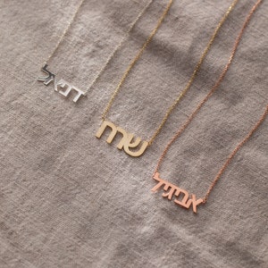 May include: Three gold necklaces with Hebrew lettering. The top necklace has the word "Shabbat" in Hebrew, the middle necklace has the word "Sha" in Hebrew, and the bottom necklace has the word "Avodah" in Hebrew.