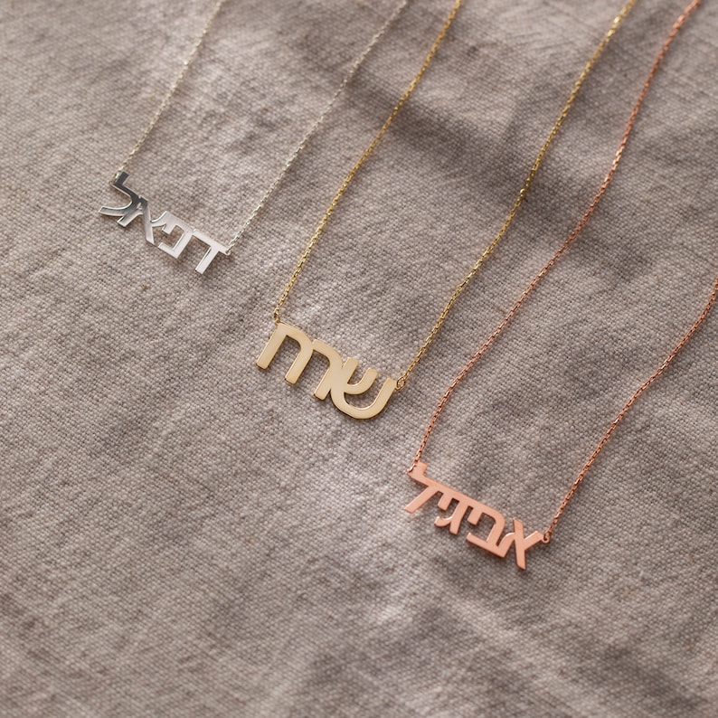 May include: Three gold necklaces with Hebrew lettering. The top necklace has the word "Shabbat" in Hebrew, the middle necklace has the word "Sha" in Hebrew, and the bottom necklace has the word "Avodah" in Hebrew.