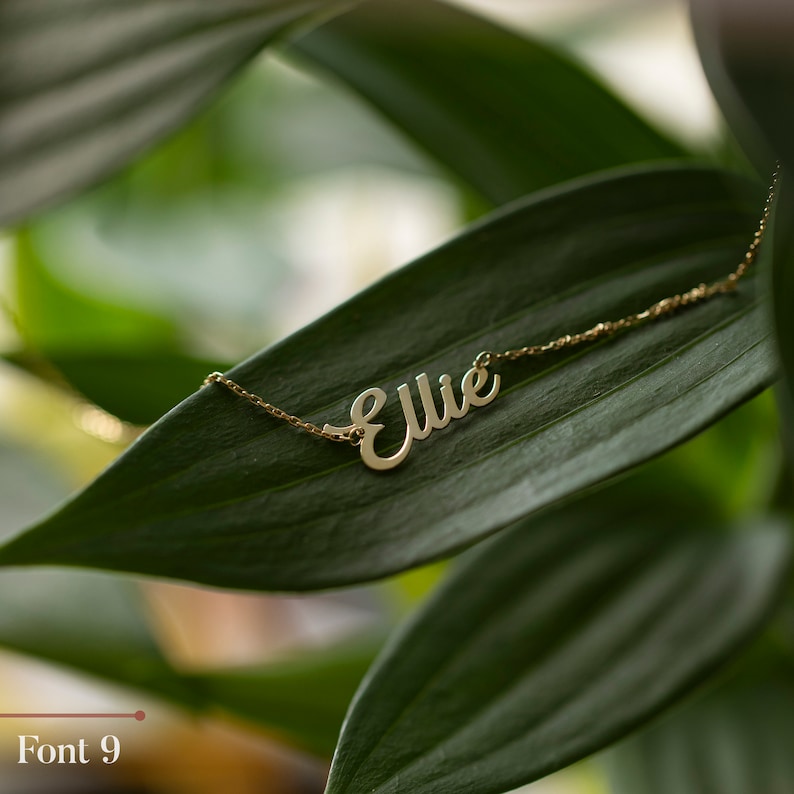 May include: Gold necklace with the name "Ellie" written in a cursive font. The necklace is on a green leaf.