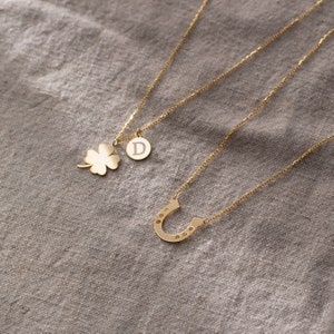 May include: Three gold necklaces with different charms. One necklace has a four-leaf clover charm, another has a horseshoe charm with small stones, and the third has a round charm with the letter D.