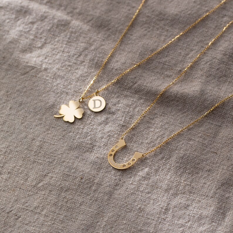 May include: Three gold necklaces with different charms. One necklace has a four-leaf clover charm, another has a horseshoe charm with small stones, and the third has a round charm with the letter D.