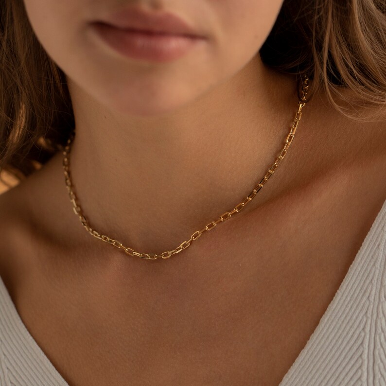 May include: A gold chain necklace with a delicate, box chain design. The necklace is worn by a person with a white shirt.