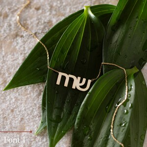 May include: A gold necklace with a Hebrew word pendant, "MY", on a green leaf background.
