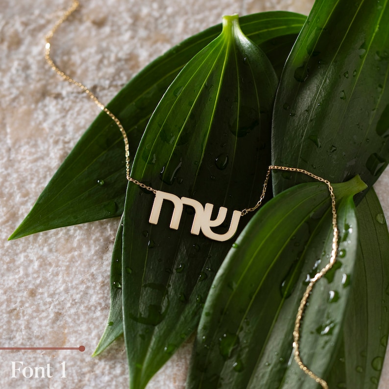 May include: A gold necklace with a Hebrew word pendant, "MY", on a green leaf background.