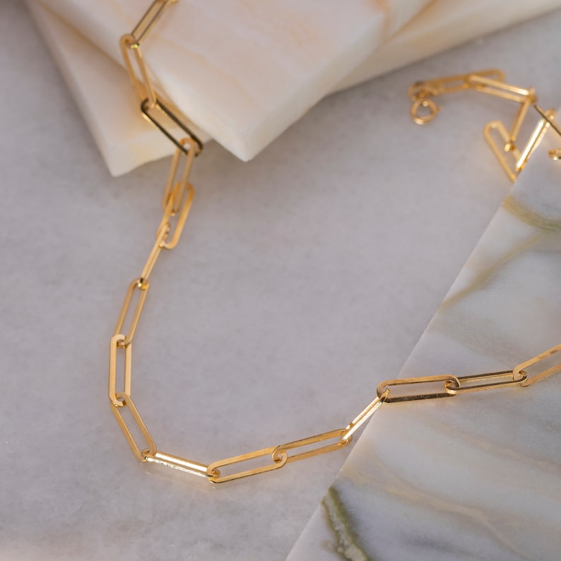 May include: A gold-colored chain necklace with rectangular links. The necklace is displayed on a white and marble-patterned surface. The chain is a simple, elegant design.