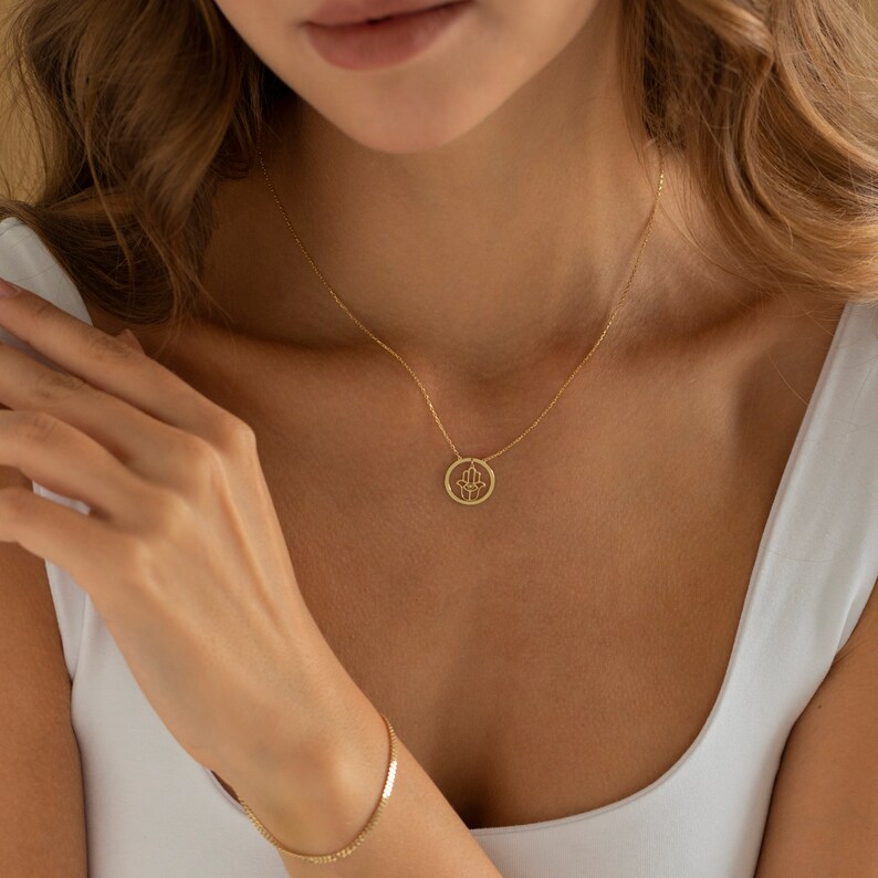 Puede incluir: Un collar de oro con un delicado colgante de mano de Hamsa. El colgante es un c&iacute;rculo con un dise&ntilde;o calado de una mano de Hamsa.