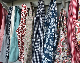 May include: A collection of 10 colorful aprons hanging on a rack. The aprons are made of different fabrics and patterns, including floral, plaid, and gingham.