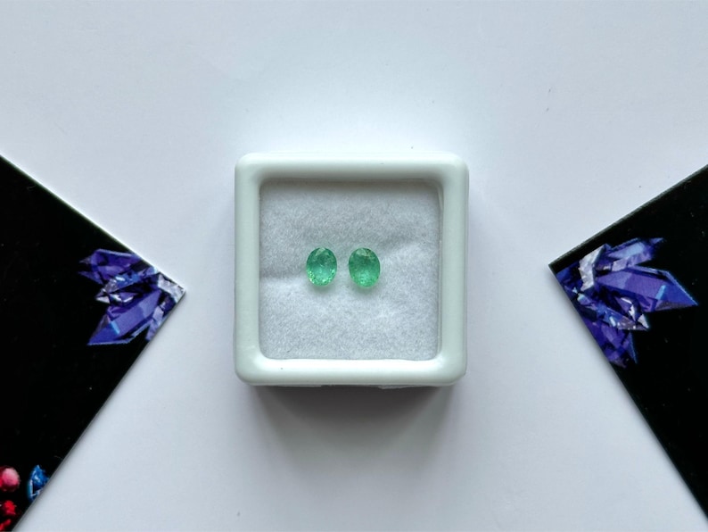 Puede incluir: Dos piedras preciosas de esmeralda ovaladas, de color verde brillante, se exhiben en una peque&ntilde;a caja de joyer&iacute;a cuadrada blanca. Las piedras preciosas est&aacute;n sobre un fondo blanco. La caja est&aacute; centrada en una superficie blanca, con tarjetas oscuras y anguladas con un dise&ntilde;o de cristal p&uacute;rpura a cada lado.