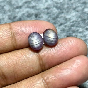 以下が含まれることがあります： ラベンダー色の2つの楕円形の磨かれた宝石。銀色の縞模様のインクルージョンがあります。滑らかで反射する表面が特徴で、手で持たれています。これらの宝石は、おそらくジュエリー用です。