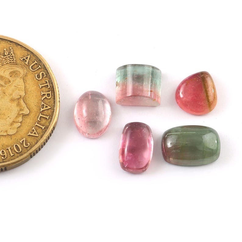 Puede incluir: Cinco piedras preciosas de diferentes formas con colores rosa, verde y blanco. Las piedras preciosas est&aacute;n todas talladas y pulidas. Se muestran sobre una superficie blanca junto a una moneda de oro australiana.