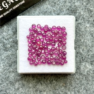 May include: A selection of small, round, polished gemstones in shades of pink and magenta, displayed in a white square box. The gemstones are scattered across the box, highlighting their colour and clarity. The box is on a grey surface.