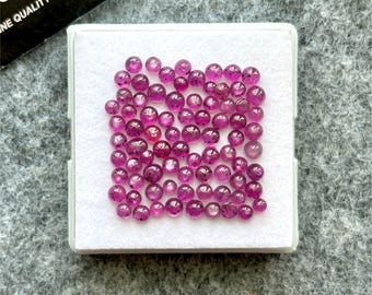 Pode incluir: Uma coleção de pequenas pedras preciosas redondas e polidas em vários tons de rosa e magenta, apresentadas numa caixa quadrada branca. As pedras preciosas estão espalhadas pela caixa, mostrando sua cor e clareza. A caixa está sobre uma superfície cinza.
