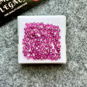 May include: A collection of small, round, polished, magenta-colored gemstones displayed in a white box. The gemstones are of varying sizes and are scattered throughout the box. The box is on a gray surface. The top left corner of the image shows a black card with white text.