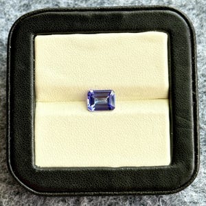 May include: A purple gemstone in a rectangular emerald cut, displayed in a black velvet ring box.
