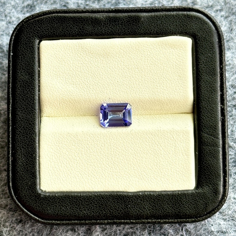 May include: A purple gemstone in a rectangular emerald cut, displayed in a black velvet ring box.