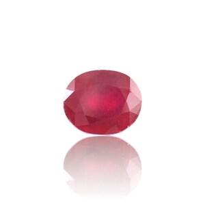 Puede incluir: Una piedra preciosa de rubí de corte ovalado con un color rojo intenso y una superficie facetada. La piedra muestra una claridad brillante y refleja la luz a través de sus bordes pulidos, exhibiendo un tono carmesí vibrante y uniforme.